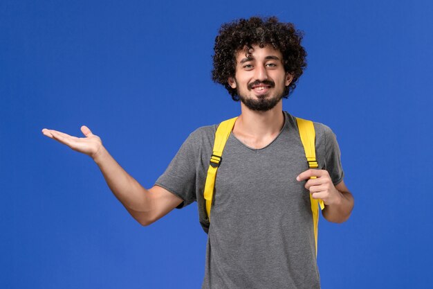 Vue de face du jeune homme en t-shirt gris portant un sac à dos jaune souriant sur le mur bleu