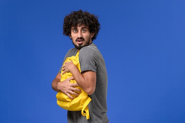 Vue de face du jeune homme en t-shirt gris tenant un sac à dos jaune sur le mur bleu