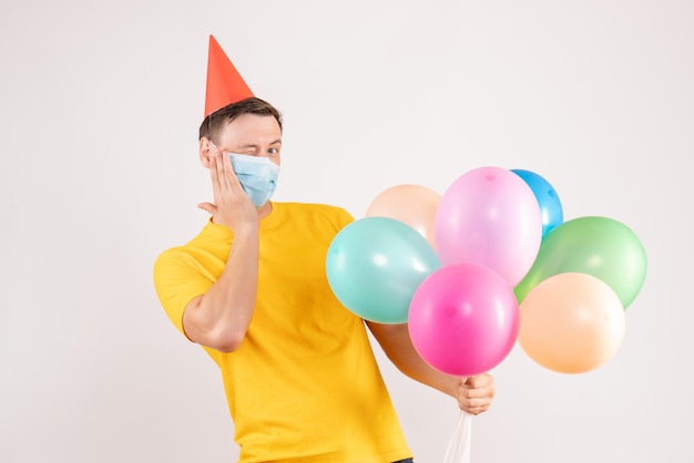 Vue de face du jeune homme tenant des ballons colorés dans un masque stérile sur un mur blanc