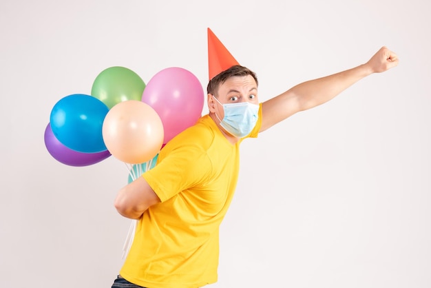 Vue de face du jeune homme tenant des ballons colorés dans un masque stérile sur un mur blanc