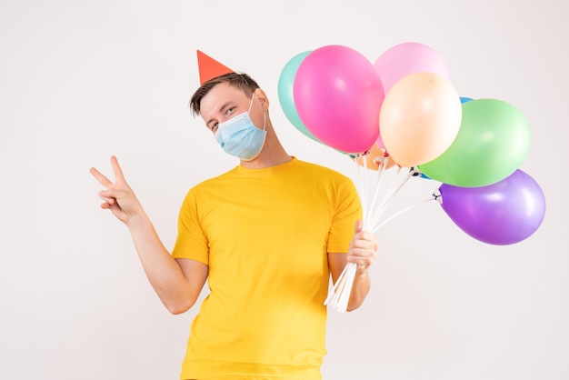 Vue de face du jeune homme tenant des ballons colorés en masque sur mur blanc