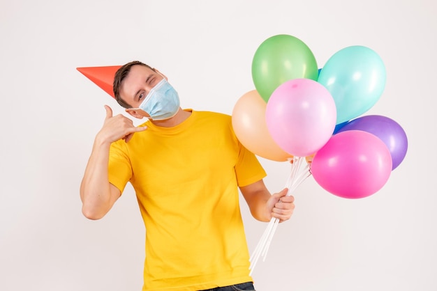 Vue de face du jeune homme tenant des ballons colorés en masque sur mur blanc