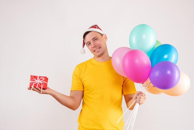 Vue de face du jeune homme tenant des ballons colorés et peu de cadeau sur un mur blanc