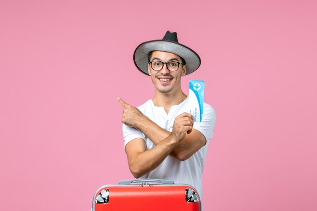 Vue de face du jeune homme tenant un billet d'avion pour des vacances sur le mur rose