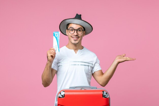 Vue de face du jeune homme tenant un billet d'avion pour des vacances sur le mur rose