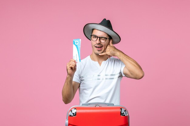 Vue de face du jeune homme tenant un billet d'avion pour des vacances sur le mur rose