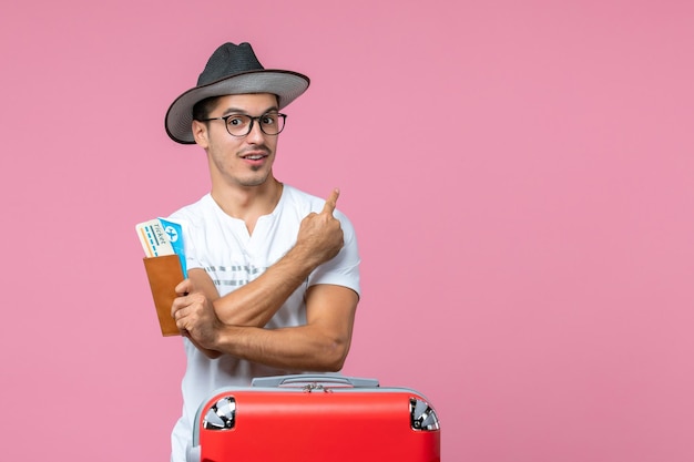 Vue de face du jeune homme tenant des billets d'avion et partant en vacances sur le mur rose