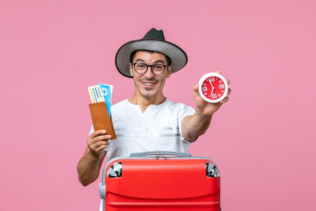 Vue de face du jeune homme tenant des billets de vacances et une horloge sur le mur rose