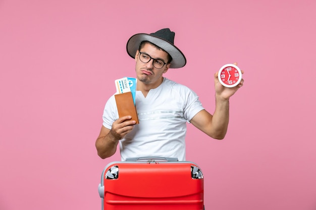 Vue De Face Du Jeune Homme Tenant Des Billets De Vacances Et Une Horloge Sur Le Mur Rose