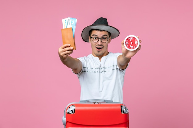 Vue de face du jeune homme tenant des billets de vacances et une horloge sur le mur rose