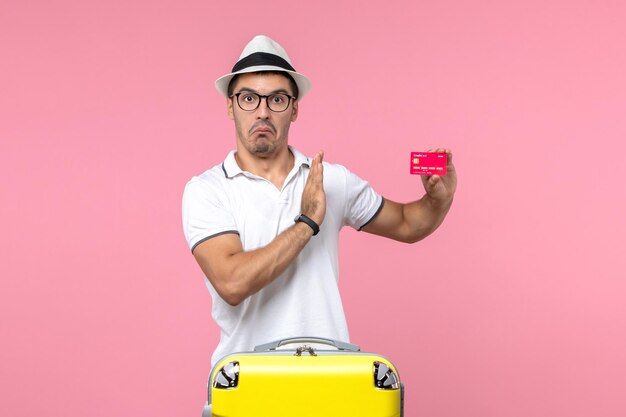 Vue de face du jeune homme tenant une carte bancaire rouge en vacances d'été sur le mur rose