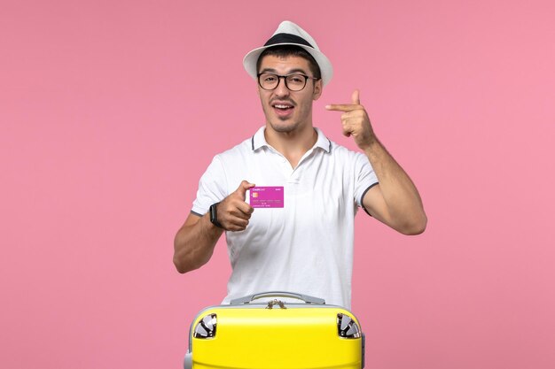 Vue de face du jeune homme tenant une carte bancaire en vacances d'été sur un mur rose clair