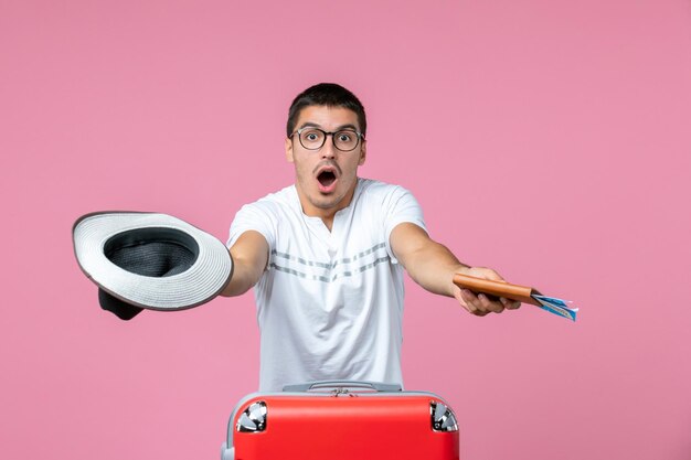 Vue de face du jeune homme tenant un chapeau et des billets d'avion sur le mur rose