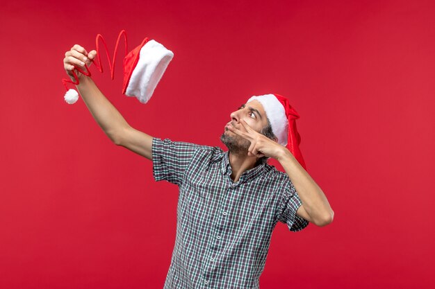 Vue de face du jeune homme tenant un chapeau de jouet de Noël sur le mur rouge