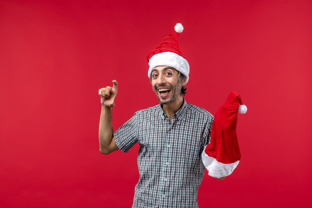 Vue de face du jeune homme tenant un chapeau de Noël rouge sur un mur rouge clair