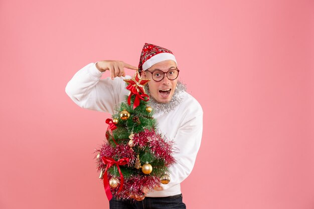 Vue de face du jeune homme tenant le petit arbre de Noël sur le mur rose