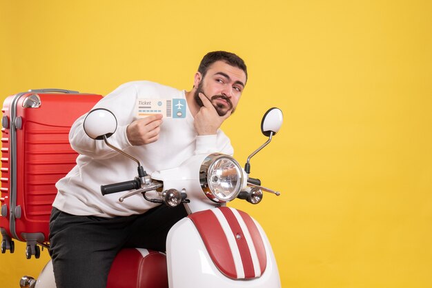 Vue de face du jeune homme voyageant pensant assis sur une moto avec une valise dessus tenant un billet sur fond jaune isolé