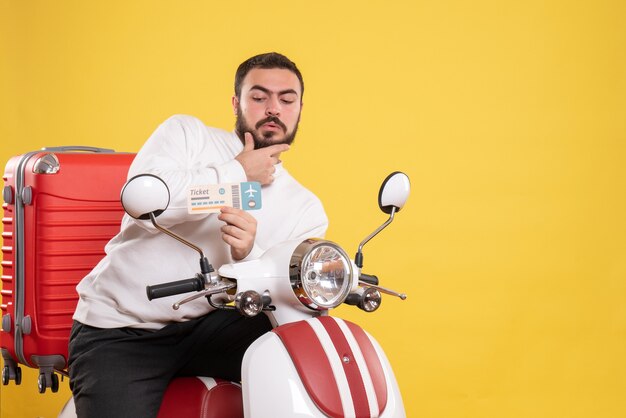 Vue de face du jeune homme voyageant réfléchi assis sur une moto avec une valise dessus tenant un billet sur fond jaune isolé