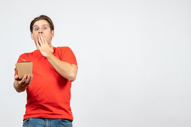 Vue de face du jeune mec émotionnel choqué en chemisier rouge tenant une petite boîte sur fond blanc