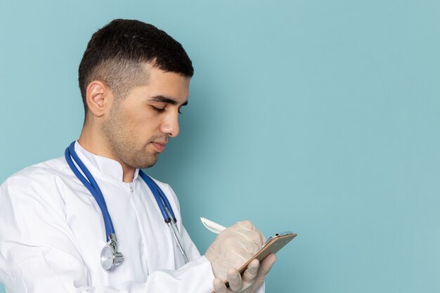 Vue de face du jeune médecin de sexe masculin en costume blanc avec stéthoscope bleu écrit des notes