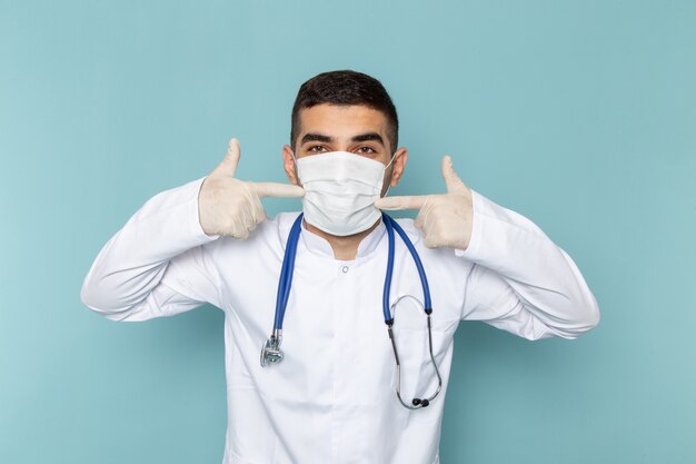 Vue de face du jeune médecin de sexe masculin en costume blanc avec stéthoscope bleu portant un masque stérile