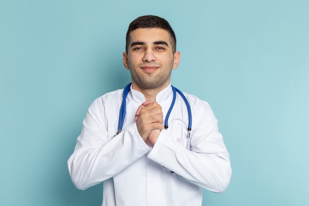 Vue de face du jeune médecin de sexe masculin en costume blanc avec stéthoscope bleu souriant posant