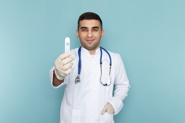 Vue de face du jeune médecin de sexe masculin en costume blanc avec stéthoscope bleu souriant et tenant le dispositif