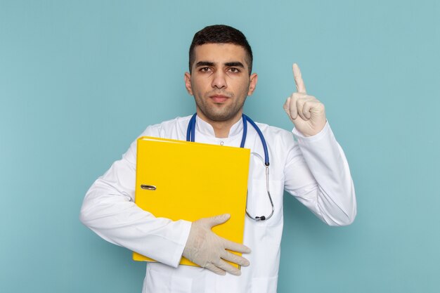 Vue de face du jeune médecin de sexe masculin en costume blanc avec stéthoscope bleu tenant des fichiers jaunes