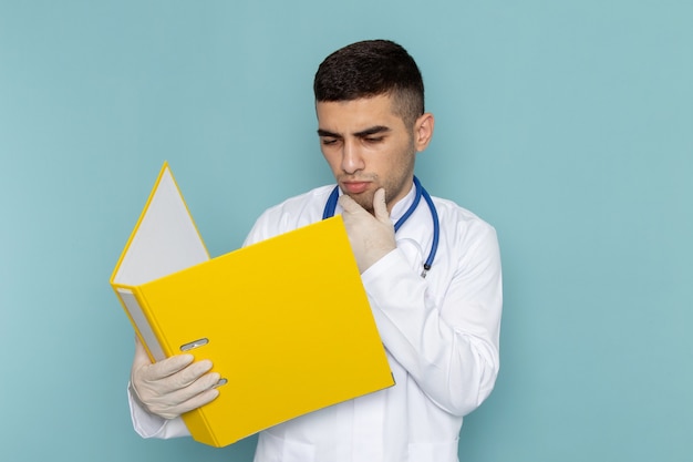 Vue de face du jeune médecin de sexe masculin en costume blanc avec stéthoscope bleu tenant la lecture de fichiers jaunes