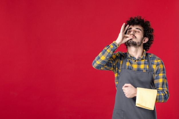 Vue de face du jeune serveur masculin souriant aux cheveux bouclés tenant une serviette faisant un geste parfait debout sur un mur rouge isolé