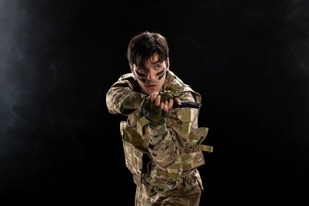 Vue de face du jeune soldat avec arme en camouflage sur mur noir