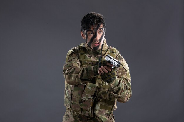 Vue de face du jeune soldat en tenue de camouflage avec arme à feu sur le mur sombre