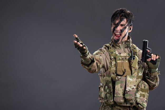 Vue de face du jeune soldat en tenue de camouflage avec arme à feu sur le mur sombre