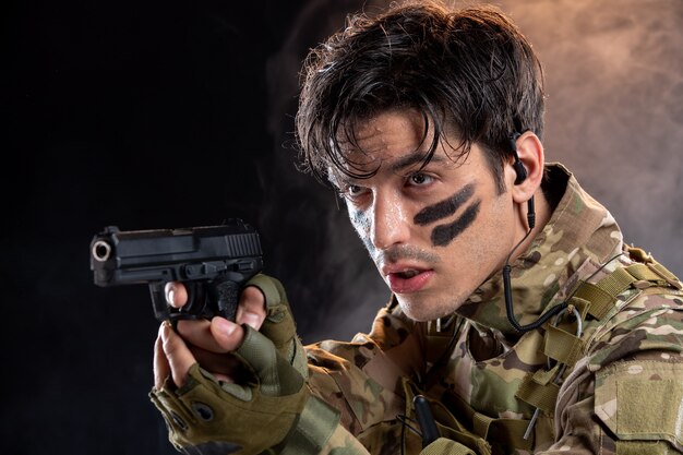 Vue de face du jeune soldat en tenue de camouflage avec pistolet sur mur noir