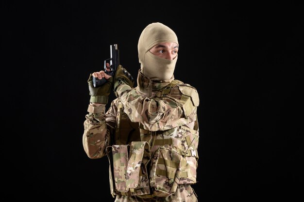 Vue de face du jeune soldat en uniforme avec arme à feu sur le mur noir