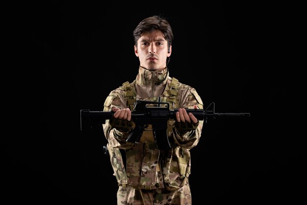 Vue de face du jeune soldat en uniforme tenant un fusil sur un mur noir