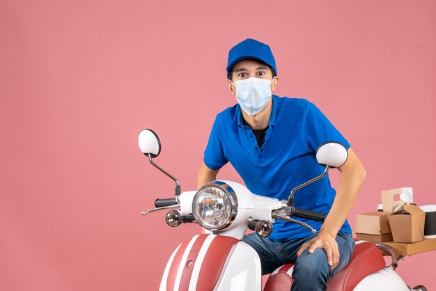 Vue de face du livreur en masque médical portant un chapeau assis sur un scooter sur fond de pêche pastel