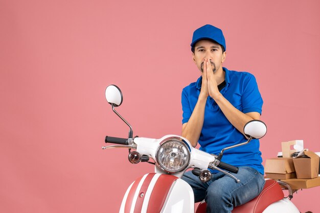 Vue de face du livreur portant un chapeau assis sur un scooter dans des pensées profondes sur fond de pêche pastel