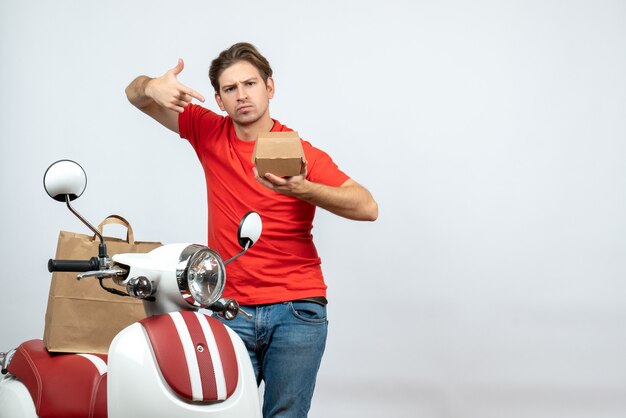 Vue de face du livreur surpris en uniforme rouge debout près de scooter pointant petite boîte sur fond blanc