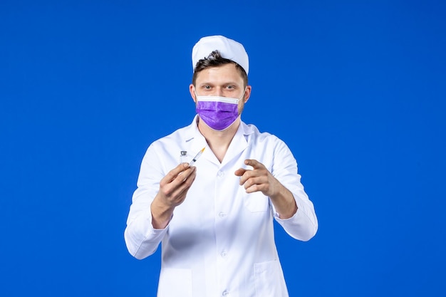 Vue de face du médecin de sexe masculin en costume médical et masque avec injection et vaccin sur bleu