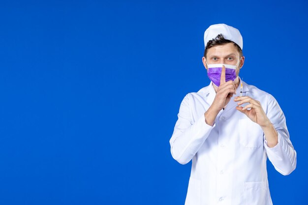 Vue de face du médecin de sexe masculin en costume médical et masque tenant le vaccin et l'injection sur bleu