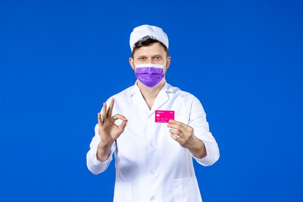 Vue de face du médecin de sexe masculin en costume médical et masque violet tenant la carte de crédit sur bleu