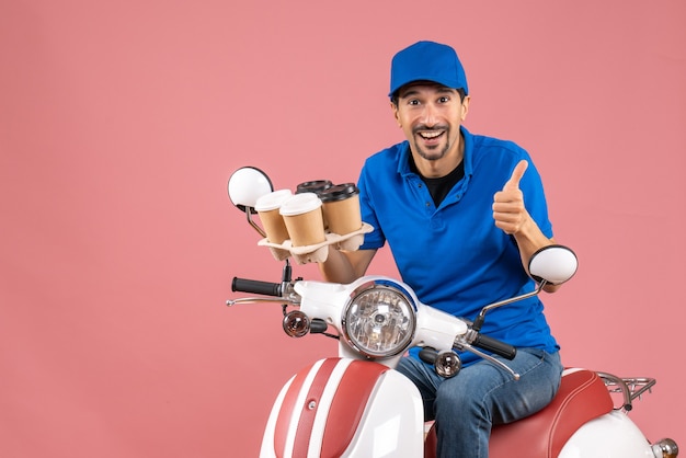 Vue de face du messager heureux positif homme portant un chapeau assis sur un scooter faisant un geste ok sur fond de pêche pastel