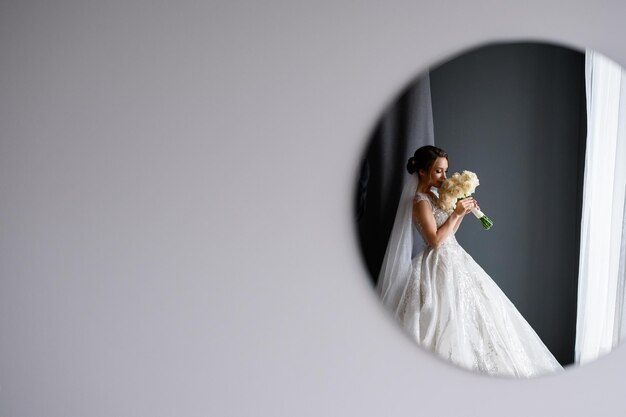 Vue de face du mur gris vide et du miroir circulaire sur lequel se reflète une jolie mariée en robe de mariée qui élève le bouquet