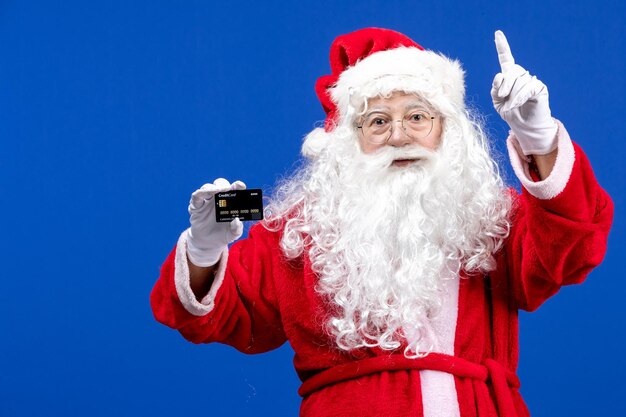 Vue de face du père noël en costume rouge tenant une carte bancaire noire sur les vacances de couleur de noël bleu présent