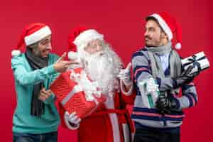 Photo gratuite vue de face du père noël avec deux hommes tenant des cadeaux sur le rouge rouge émotion cadeau du nouvel an noël