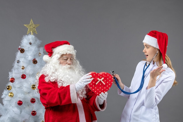 Vue de face du père Noël avec femme médecin qui observe présent avec stéthoscope sur le mur gris