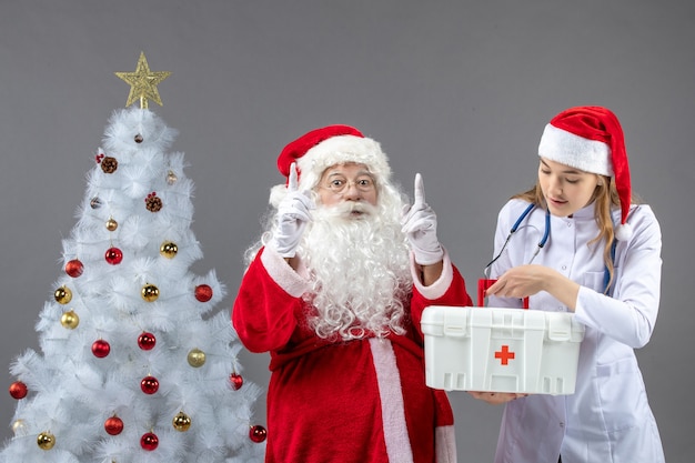 Vue De Face Du Père Noël Avec Femme Médecin Qui Tient Une Trousse De Premiers Soins Sur Le Mur Gris