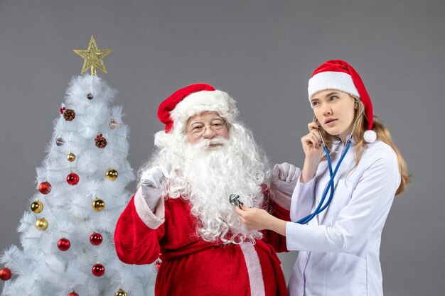 Vue de face du père Noël avec femme médecin qui vérifie son état de santé sur un mur gris