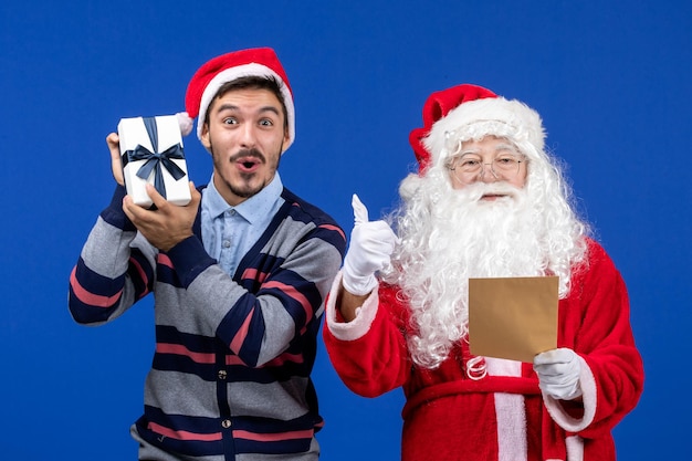 Vue de face du père noël avec un jeune homme lisant une lettre et tenant un présent sur le mur bleu